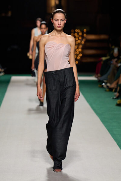 Modelka na pokazie Victoria Beckham Spring 2025 w wieczorowym looku łączącym romantyczne body w kolorze nude z przeskalowanymi, ciemnymi spodniami o nietypowej, asymetrycznej talii. Stylizacja inspirowana architekturą i geometrią mody.