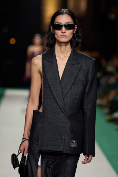 Modelka prezentuje awangardowy garnitur z kolekcji Victoria Beckham Spring 2025 – marynarka z asymetrycznym wycięciem i geometrycznymi liniami połączona z przeskalowanymi spodniami. Stylizacja wpisuje się w trend redefinicji klasyki.