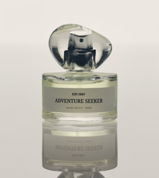 H&M zamienniki drogich perfum: Elegancki flakon perfum o nazwie „Adventure Seeker” z napisem „EST. 1947” i adnotacją „Eau de Toilette Vegan”, stojący na lustrzanej powierzchni, odbijającej jego kształt. Przezroczysta butelka z zielonkawym płynem oraz fantazyjnie uformowanym, rzeźbiarskim korkiem z przezroczystego szkła nadaje całości nowoczesny i wyrafinowany charakter.