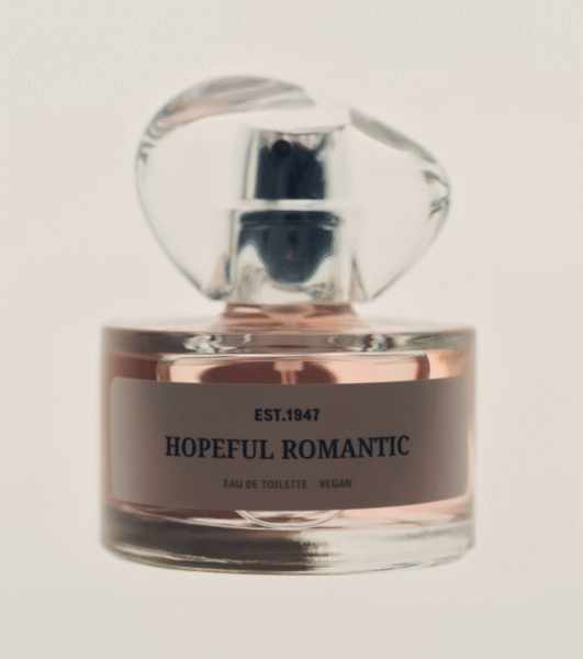 Perfumy „Hopeful Romantic” w subtelnie różowym odcieniu, umieszczone w eleganckim, przezroczystym flakonie z designerskim korkiem. Na etykiecie widnieje napis „EST. 1947”, a także informacja o wegańskim składzie. Całość ukazana na jasnym tle, tworząc romantyczną i czystą atmosferę.