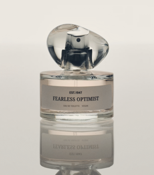Flakon perfum „Fearless Optimist” z jasnoróżowym płynem, odbijający się w lustrzanej powierzchni. Przezroczysta butelka z zaokrąglonym, szklanym korkiem i minimalistyczną etykietą z napisem „EST. 1947”. Kompozycja utrzymana w eleganckim, spokojnym stylu.