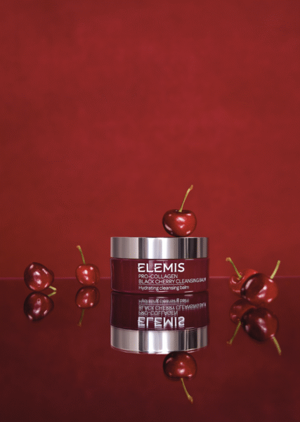 Artystyczna kompozycja balsamu oczyszczającego do twarzy Elemis z edycji Black Cherry. Produkt w bordowym słoiczku odbija się w lustrzanej powierzchni, otoczony świeżymi wiśniami na intensywnie czerwonym tle, podkreślającym nawilżające i odżywcze właściwości kosmetyku. Kosmetyczne FOMO na czerwiec 2025