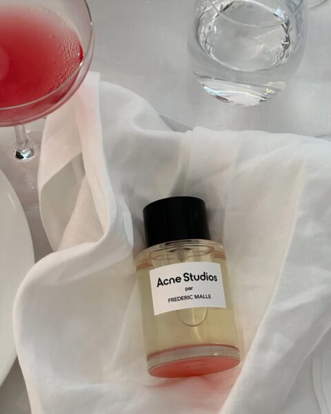 Flakon perfum Acne Studios par Frédéric Malle ustawiony na białej, lekko pogniecionej tkaninie, obok kieliszka z różowym napojem oraz szklanki z wodą. Perfumy mają prostą, przezroczystą butelkę z czarnym korkiem i minimalistyczną etykietą. Kompozycja nawiązuje do estetyki francuskiego stołu, łącząc nowoczesny design z subtelną elegancją. H&M zamienniki drogich perfum.