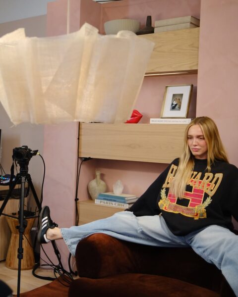 Alexandra Cooper, autorka podcastu Call Her Daddy. Na zdjęciu siedzi z rozkraczonymi nogami w studiu. Ma na sobie jesny, bluzę z nadrukiem i sneakersy adidas.