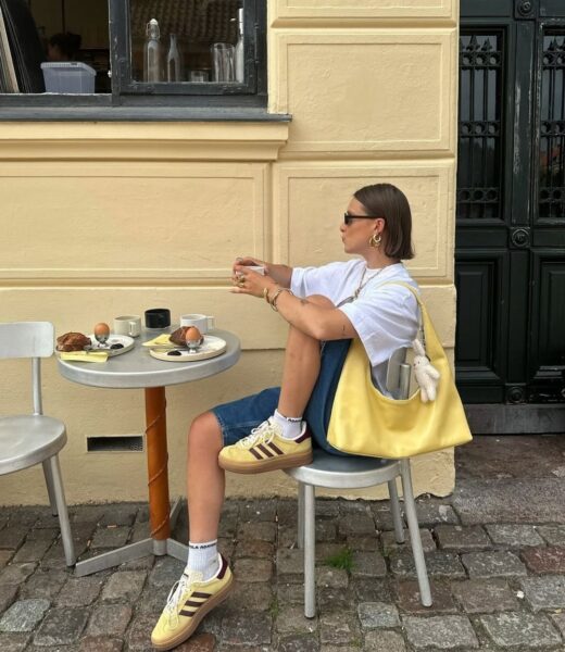 Kobieta w krótkich ciemnych włosach siedzi przy stoliku przy budynku. Nosi biały luźny T-shirt, jeansowe spodnie do kolan oraz żółte sneakersy z kontrastującymi paskami. Na jej ramieniu wisi duża żółta torebka. Kobieta właśnie podnosi filiżankę.