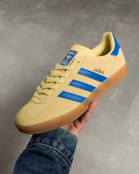 Modne seakersy Adidas Gazelle w odcieniu maślanej żółci na lato 2025. Sneakersy mają niebieskie detale – paski, zapiętek i naszywkę na języku. Są trzymane przez osobę noszącą jeansową koszulę lub kurtkę.