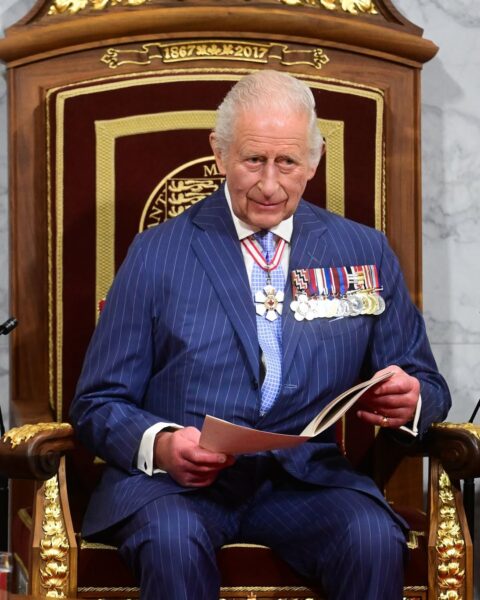 Król Karol III siedzi na królewskim tronie w prążkowanym garniturze i z licznymi odznaczeniami – podczas oficjalnej ceremonii.