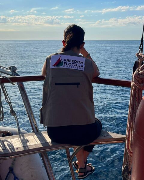 Greta Thunberg Gaza: Kobieta siedzi na ławce na pokładzie żaglówki, z widokiem na spokojne morze. Ma na sobie bezrękawnik z logo „Freedom Flotilla Coalition” — organizacji znanej z protestów morskich. Ujęcie skupia się na jej plecach i patrzeniu w stronę horyzontu, co buduje refleksyjny nastrój.