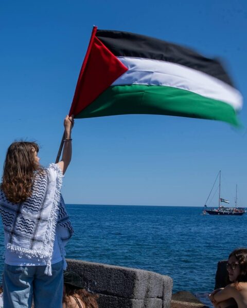 Dziewczyna odwrócona tyłem stoi na nabrzeżu, trzymając wysoko rozwiniętą flagę Palestyny. Ma na sobie białą koszulkę i keffiyeh. Obok siedzą inne osoby, a w tle na morzu widoczny jest jacht z taką samą flagą. Zdjęcie oddaje atmosferę manifestacji solidarnościowej.