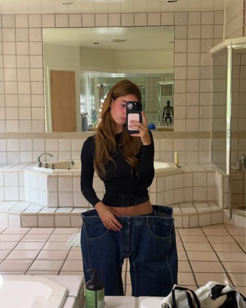 Selfie influencerki w łazience – prezentuje stylizację z viralowymi square jeans o sztywnej, przerysowanej formie. Zestawienie z dopasowaną czarną bluzką podkreśla kontrast pomiędzy górą a dolną częścią outfitu.