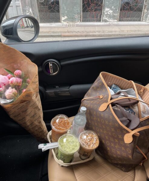 Siedzenie pasażera z torbą Louis Vuitton, bukietem kwiatów, trzema kawami na zimno i jedną zieloną herbatą matcha w uchwycie.