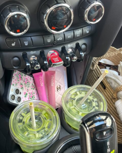 Wnętrze samochodu z dwoma zielonymi smoothie w uchwytach, telefonem z kwiecistym etui, kosmetykami Rhode i portfelem Chanel.