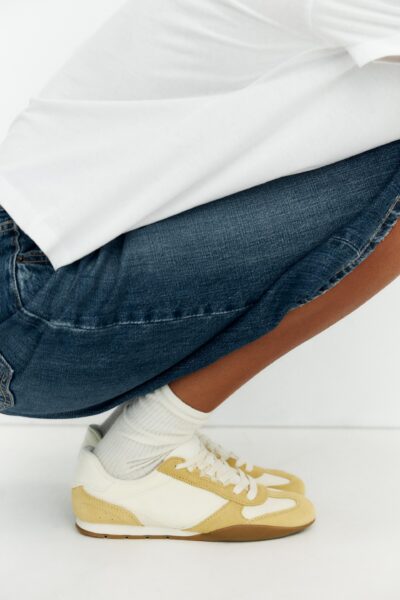 Zdjęcie produktowe modnych sneakersów w kolorze butter yellow na lato 2025. Kobieta na fotografii nosi je do białych skarpetek, jeansowych spodni i białej koszulki.